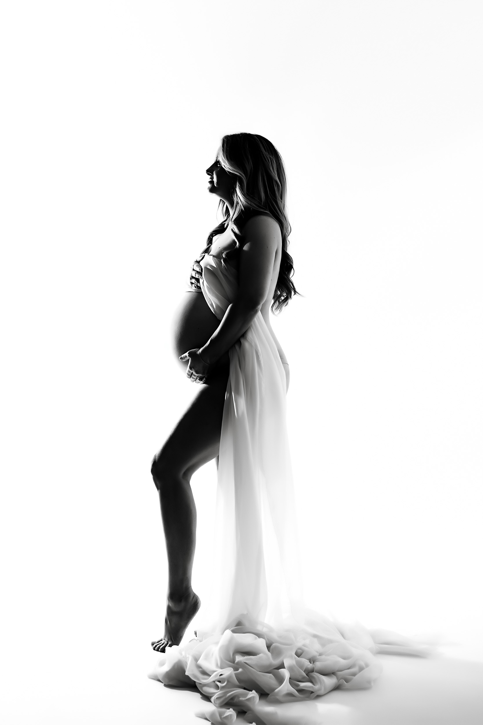 A Pregnant Woman Stands In Profile, Draped In Flowing Sheer Fabric, During A Maternity Session Against A White Background In This Black And White Photo.
