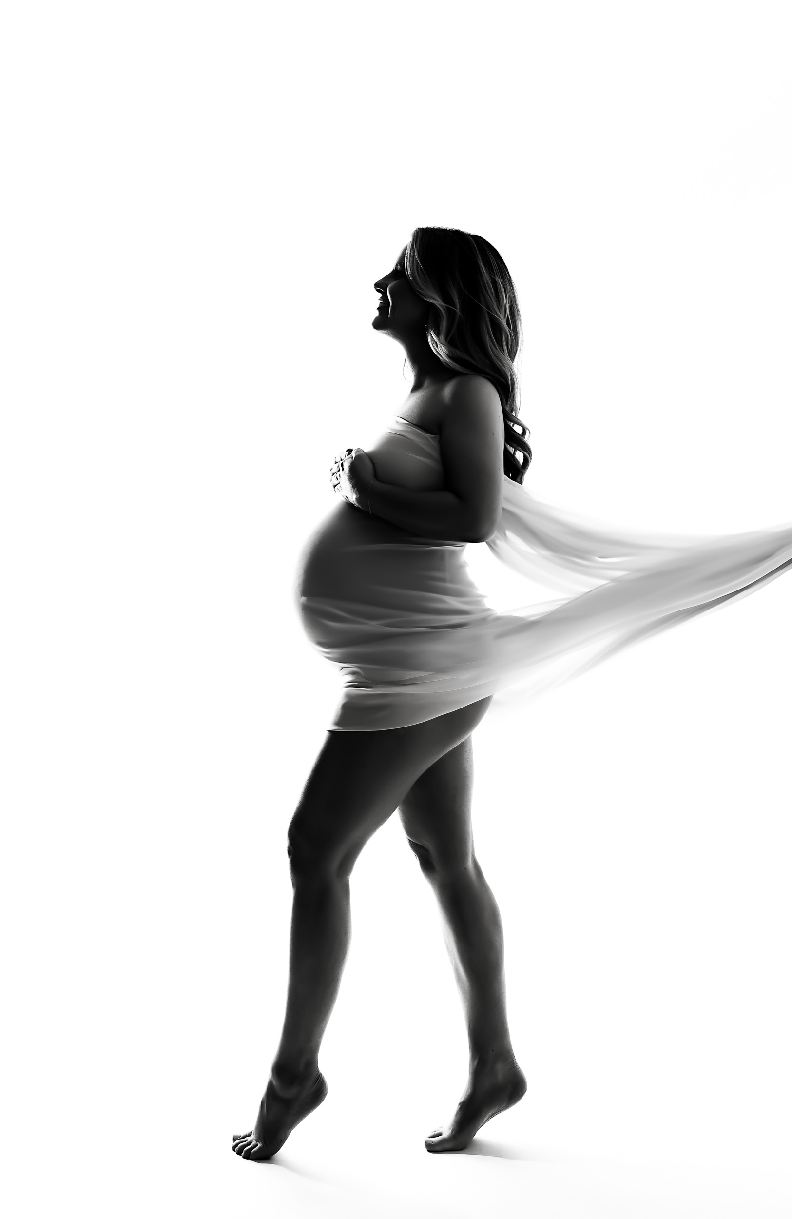 Black And White Image From A Maternity Session, Capturing A Pregnant Woman In Profile, Standing Barefoot And Draped In Flowing Fabric Against A White Background.