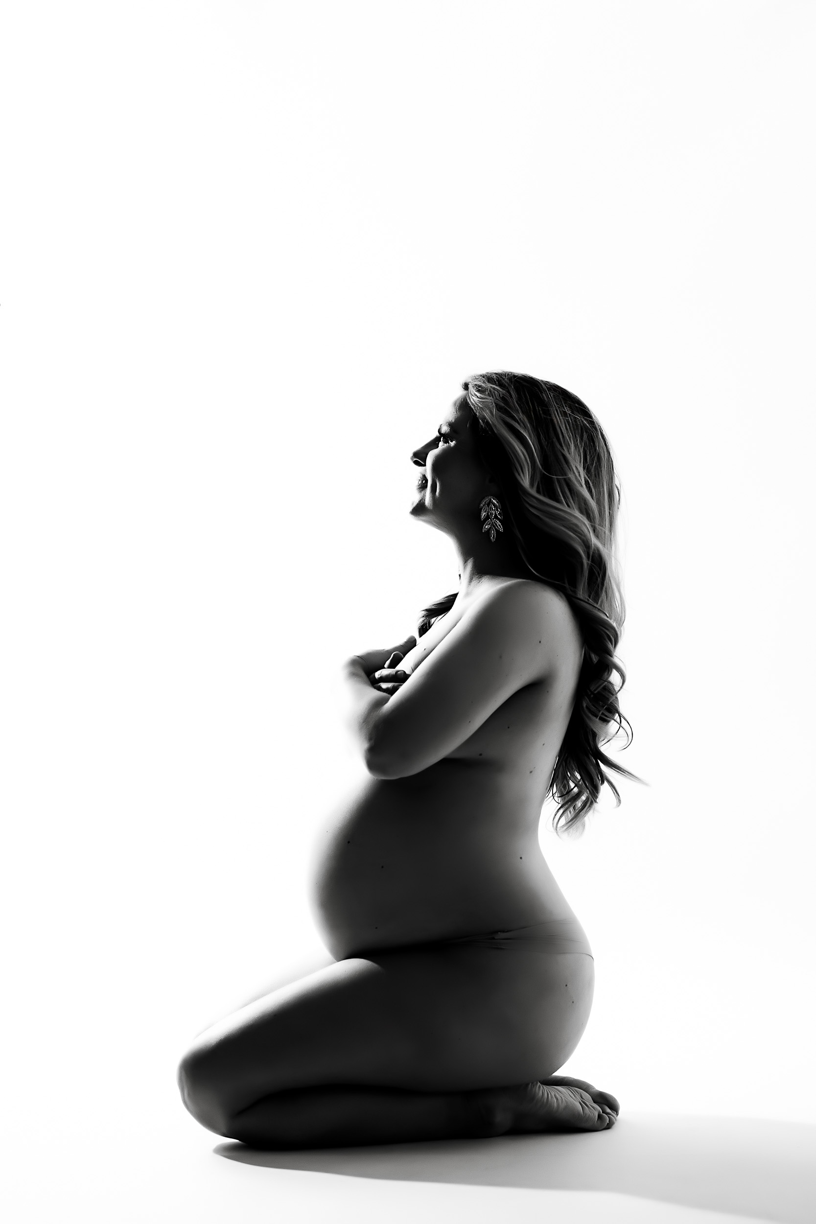 Black And White Photo From A Maternity Session, Showing A Pregnant Woman Kneeling Sideways, Covering Her Chest With Her Arms, And Looking Forward.