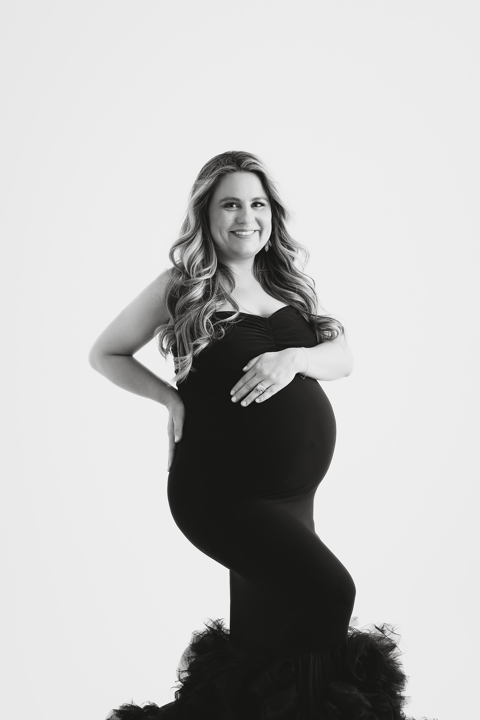 A Pregnant Woman In A Fitted, Strapless Black Dress Poses And Smiles At The Camera Against A Plain, Light Background, Capturing A Radiant Moment During Her Maternity Session.