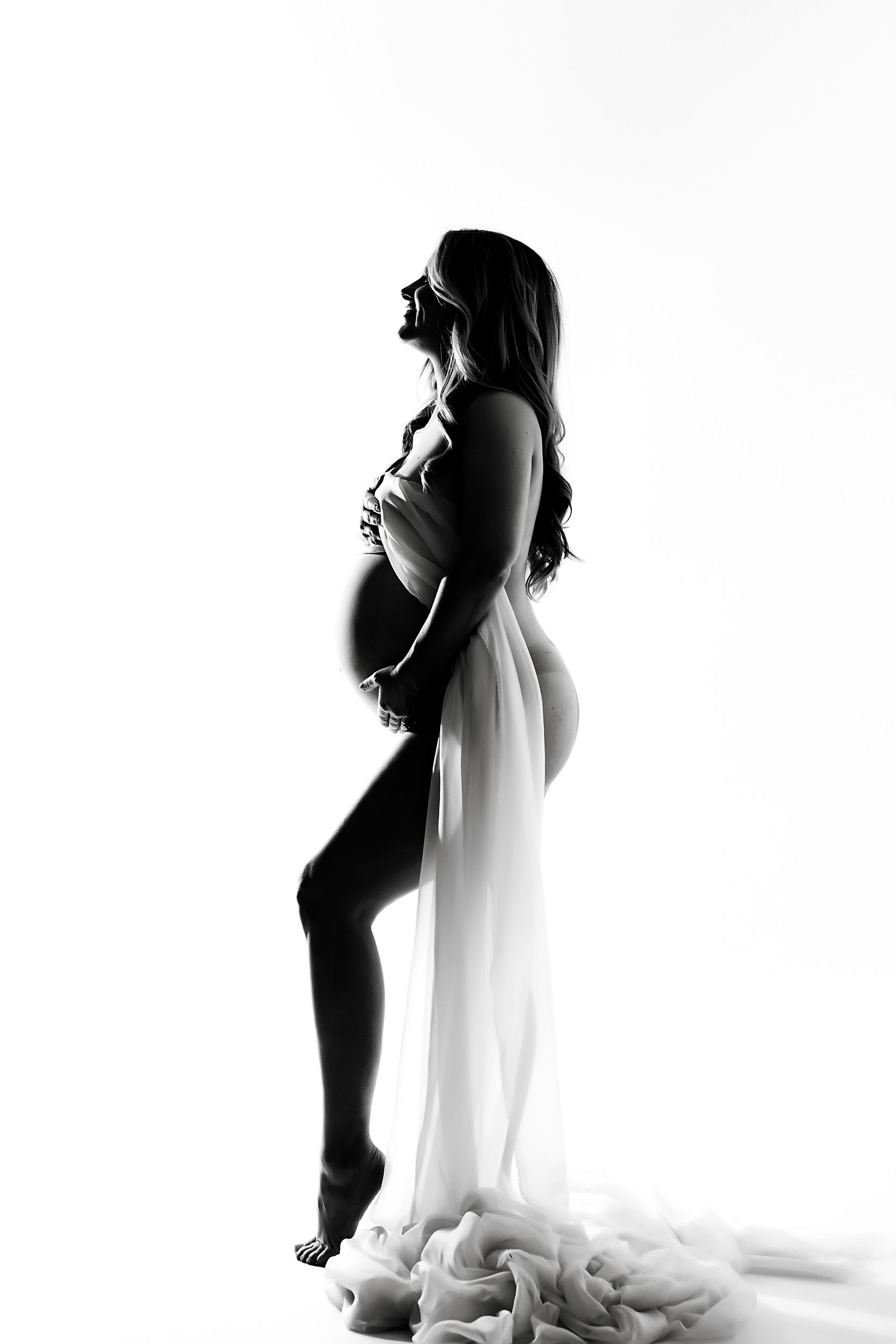 Black And White Photo Of A Pregnant Woman In Profile, Draped In Sheer Fabric, Standing Barefoot Against A White Background—An Intimate Maternity Session Capturing The Beauty Of Motherhood.
