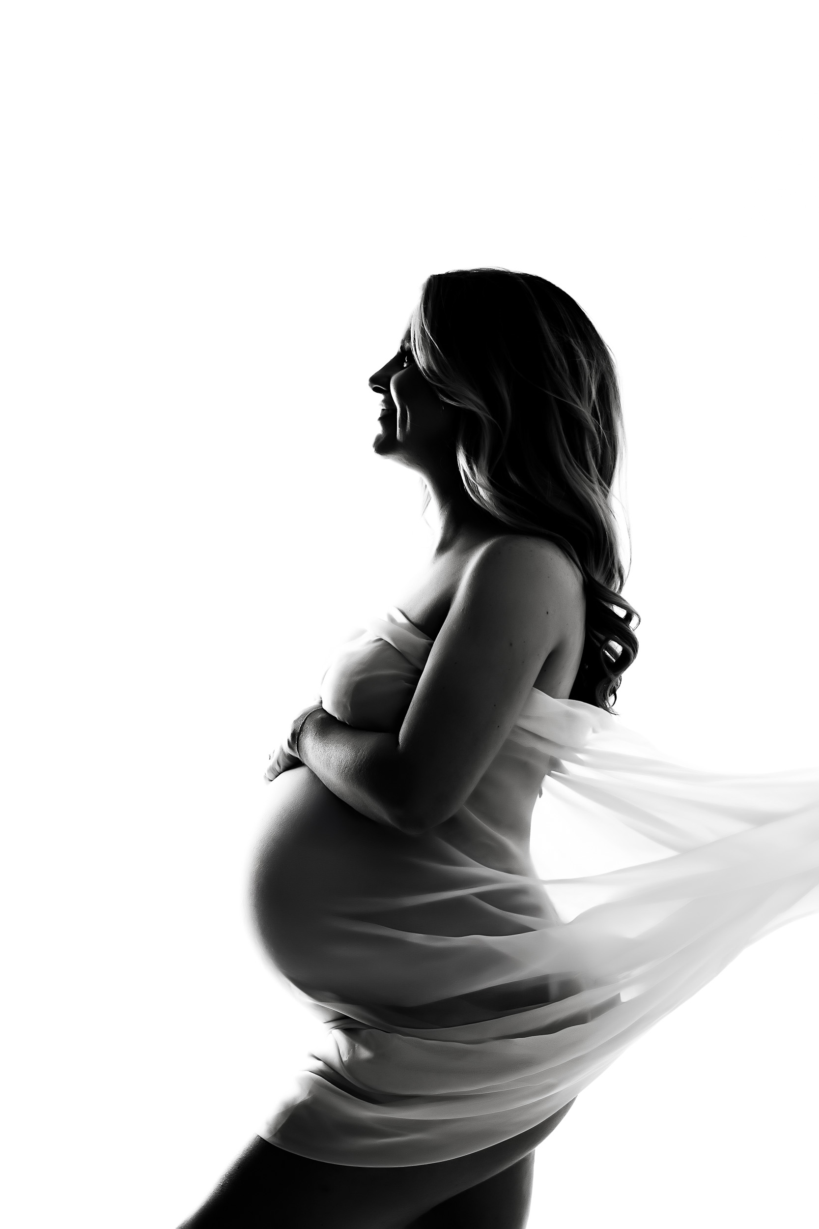 Black And White Side Profile Of A Pregnant Woman Draped In Sheer Fabric, Standing Against A White Background—An Elegant Portrait Perfect For A Maternity Session.