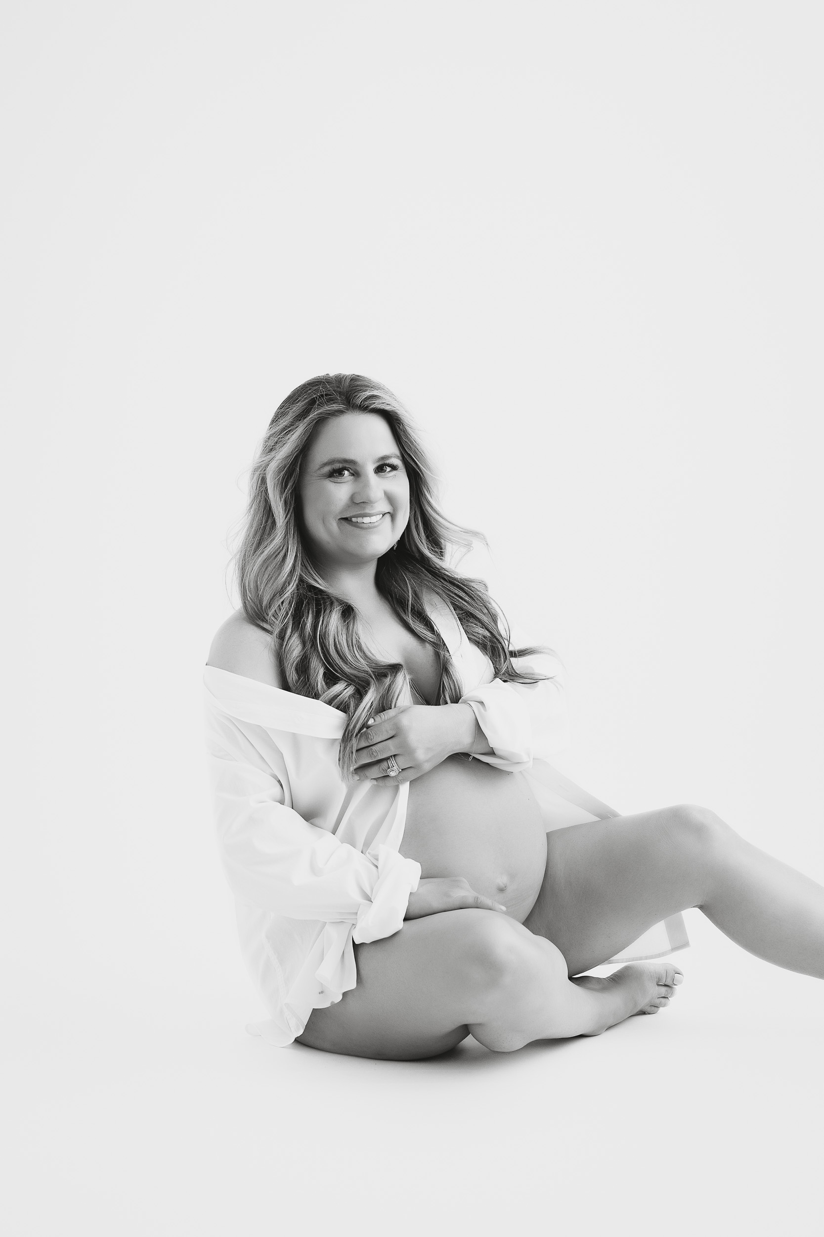 A Pregnant Woman Sits On The Floor In A White Open Shirt, Smiling At The Camera, With One Hand Resting On Her Exposed Belly During A Black And White Maternity Session.