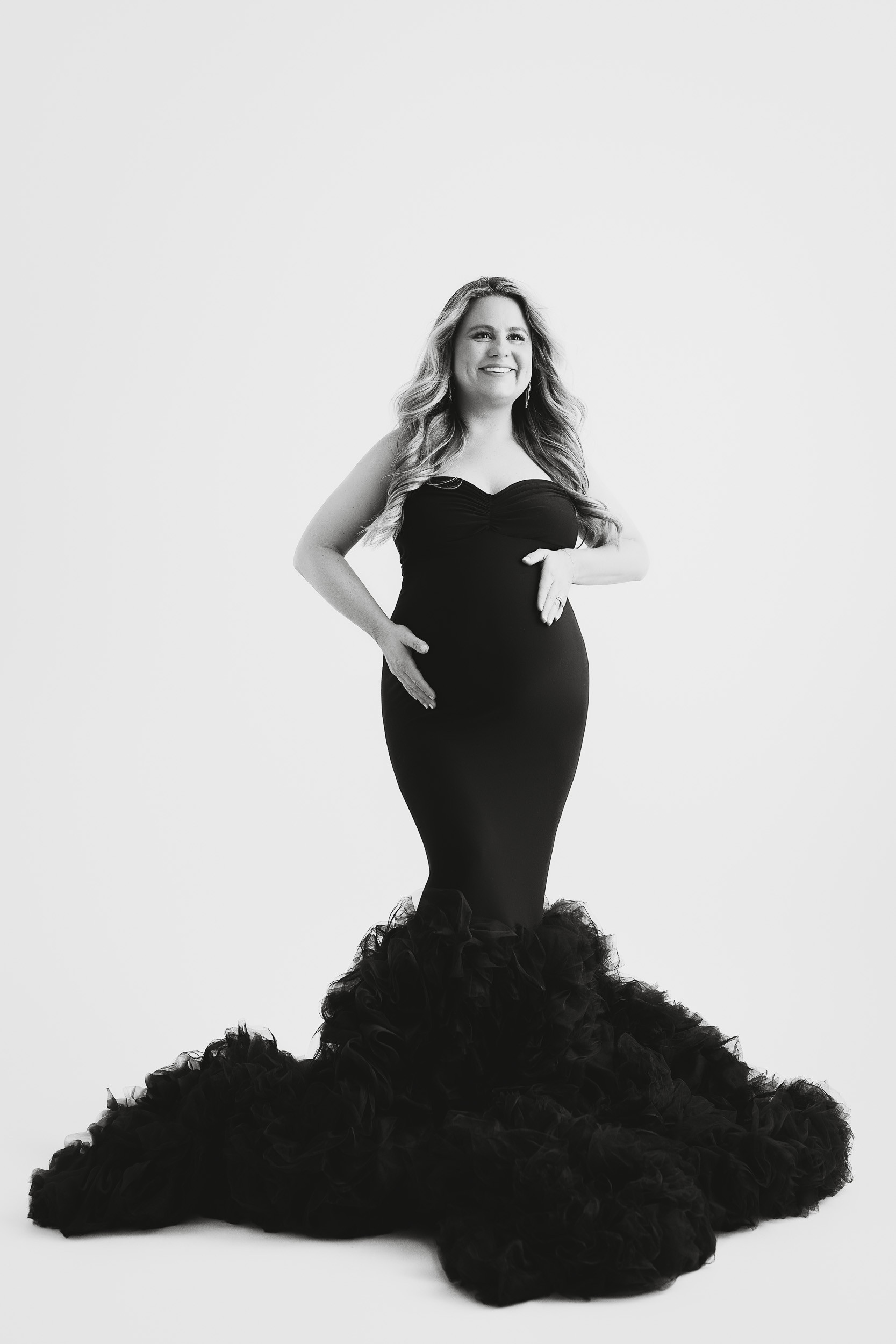 A Woman In A Fitted, Strapless Black Gown With A Ruffled Train Poses Against A White Background During Her Maternity Session, Resting Her Hands On Her Baby Bump And Smiling.