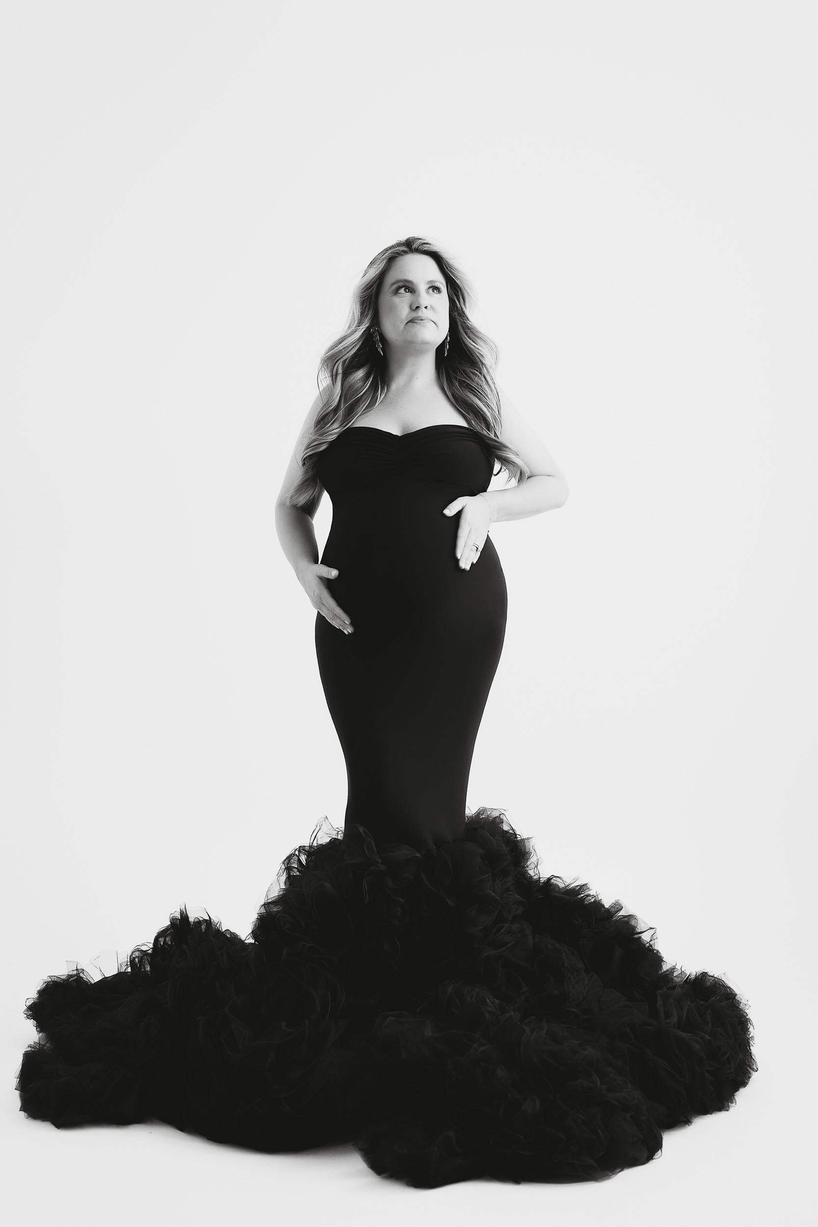 A Pregnant Woman In A Strapless, Form-Fitting Black Gown With A Ruffled Train Stands Against A White Background, Looking Slightly Upward—Capturing The Elegance And Anticipation Of Her Maternity Session.
