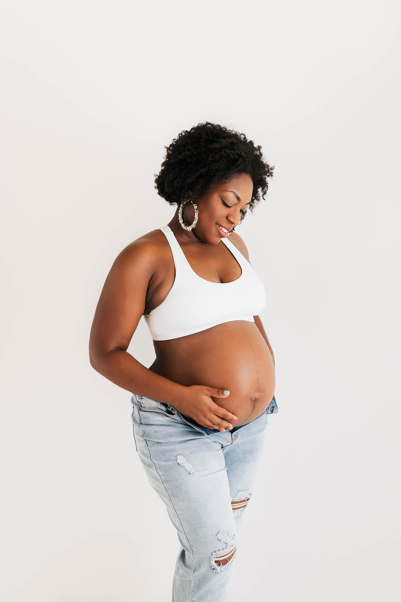 Pregnant Woman Smiling And Holding Baby Bump In White Top And Jeans During Studio Dallas Maternity Session