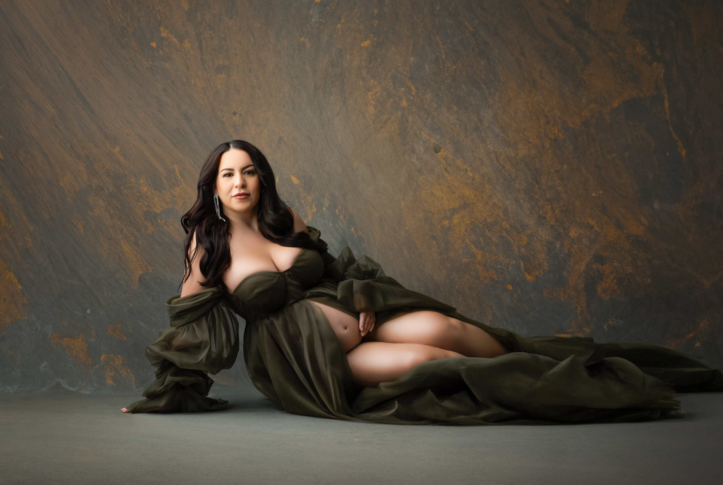 Creative Pregnancy Photos Of A Pregnant Woman In An Elegant Green Gown, Reclining Against A Textured Brown And Gray Backdrop, Captured By A Dallas Maternity Photographer