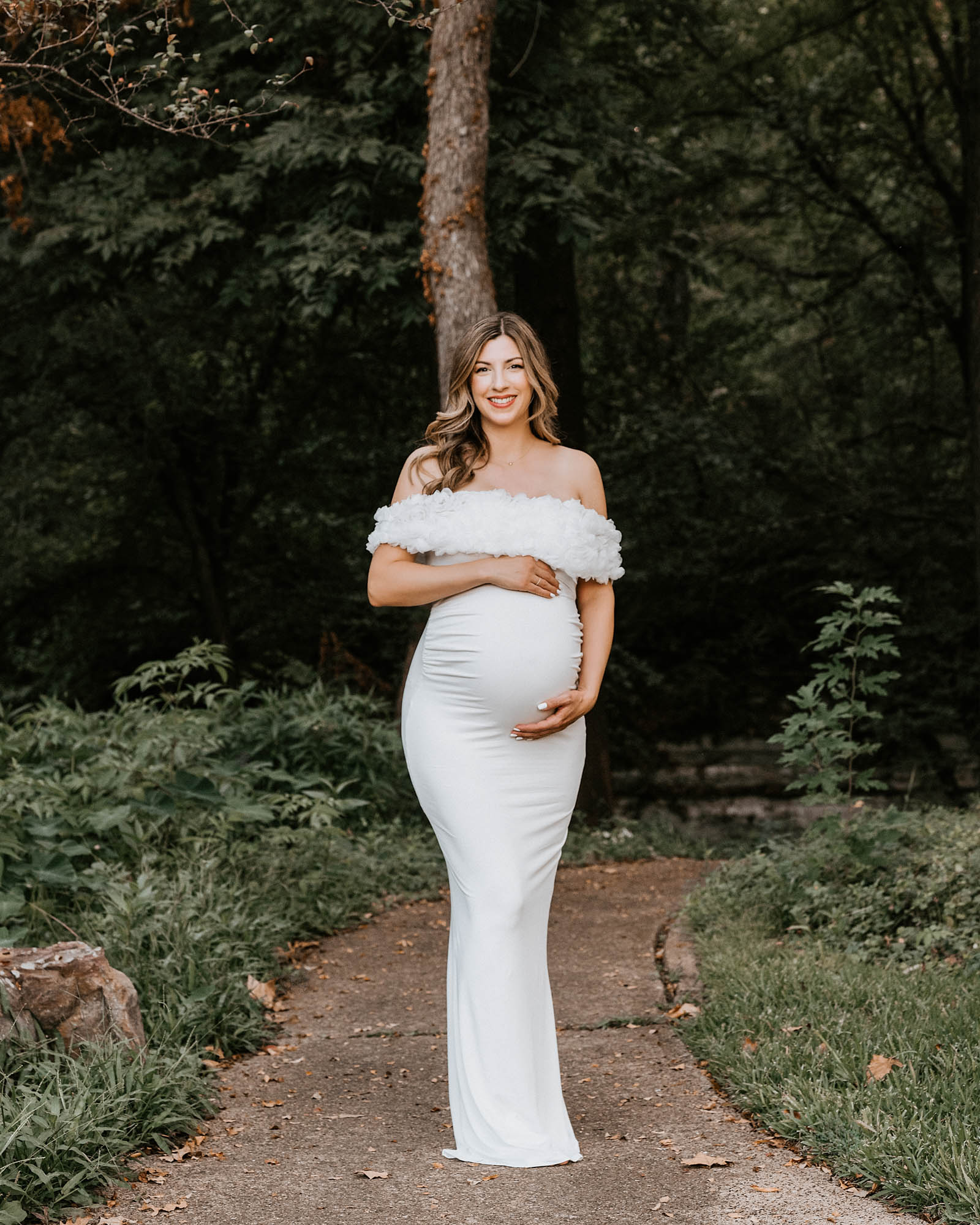 Pregnant Woman In Elegant White Gown Holding Belly During Outdoor Maternity Photoshoot In Dallas