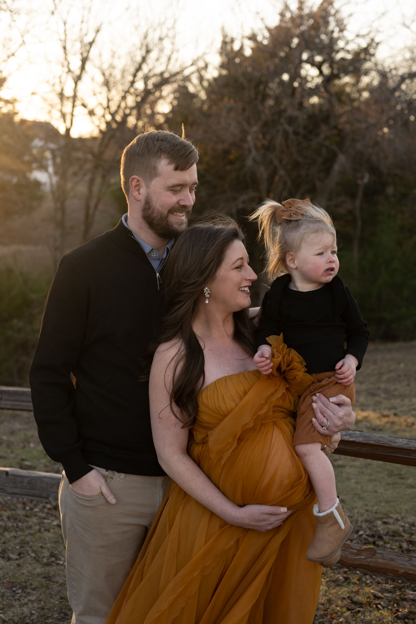 Smiling family during outdoor maternity photoshoot in Dallas, mother in flowing mustard dress holding child