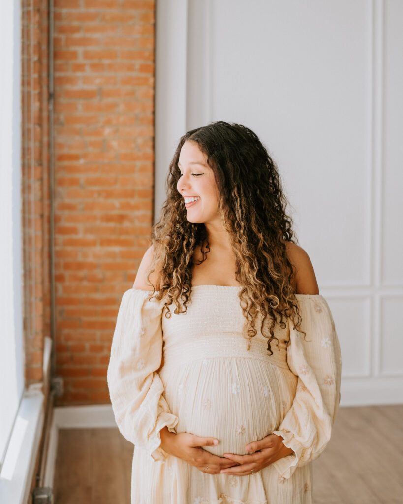 Pregnant Woman Smiling While Holding Belly Near Window During Dallas Maternity Shoot