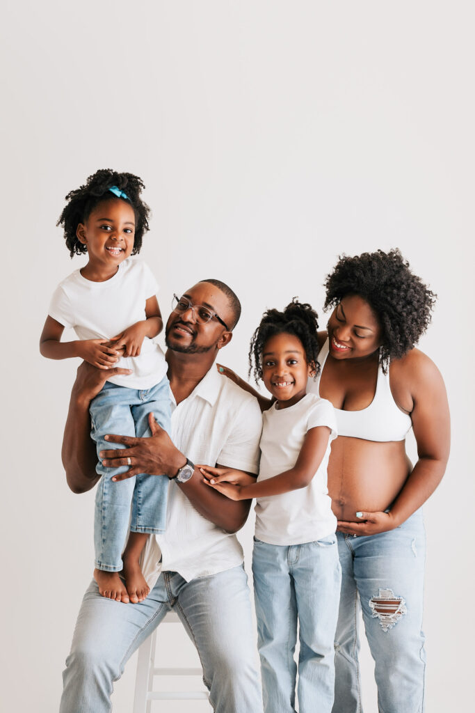 Best Maternity Poses For Natural And Elegant Portraits 10 Joyful Family Maternity Portrait In Dallas Featuring A Pregnant Mother, Father, And Two Young Daughters Wearing Matching White Tops And Jeans – Maternity Photography In Dallas