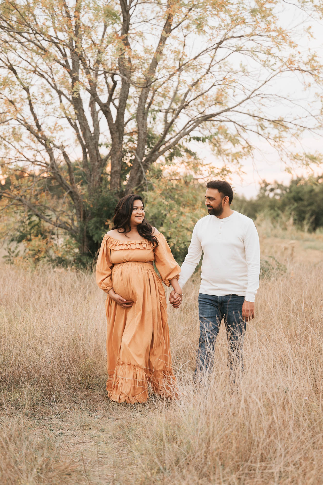 Pregnant Mother And Partner Holding Hands While Walking Through A Natural Field For A Pregnancy Session In Dallas