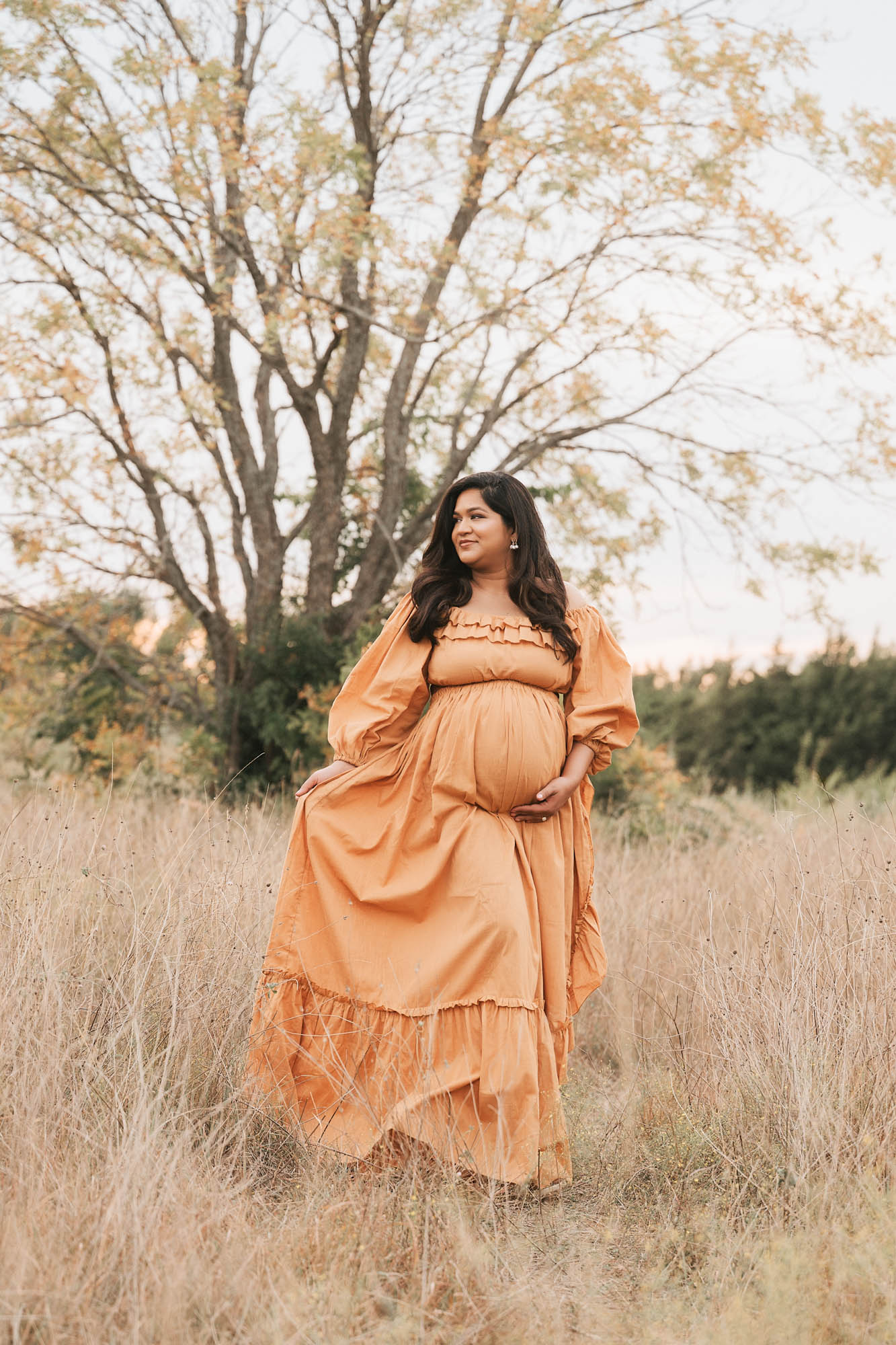 Expecting Mother In A Flowing Mustard Gown Walking Through Tall Grass During An Outdoor Maternity Session In Dallas