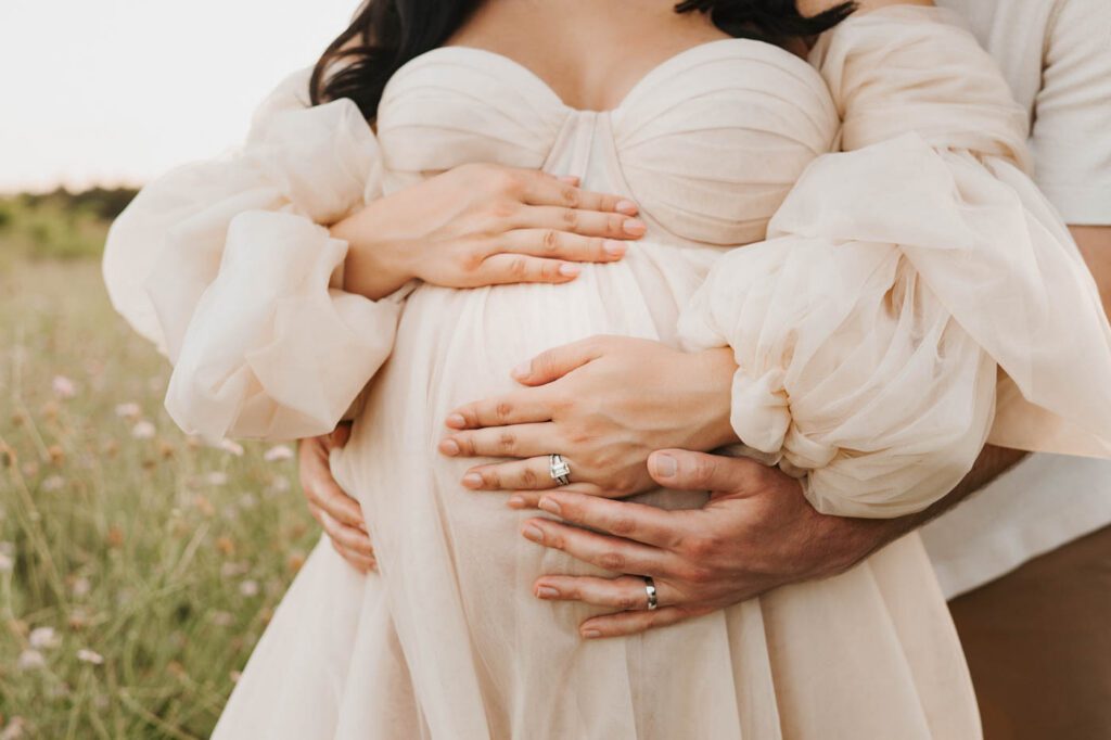 Close-Up Of Expecting Parents' Hands Resting On A Baby Bump During An Outdoor Maternity Session.