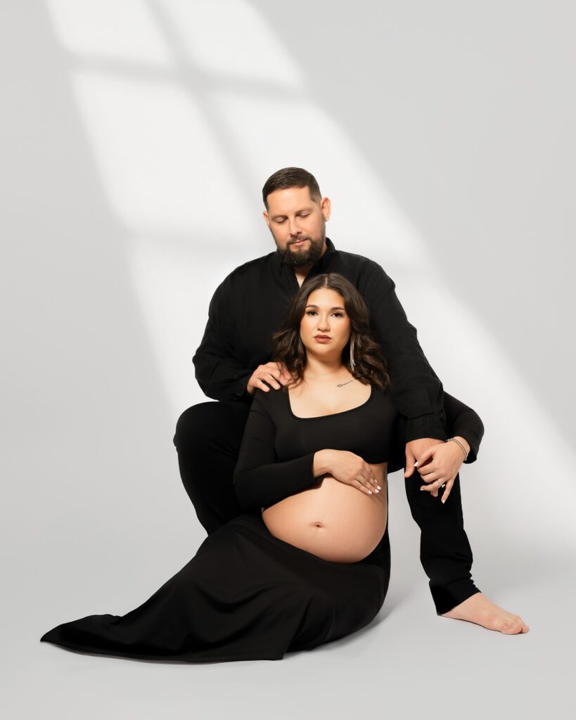 Dallas Maternity Photography Portrait Of An Expectant Couple Posed Together In Coordinated Black Clothing On A White Studio Background.