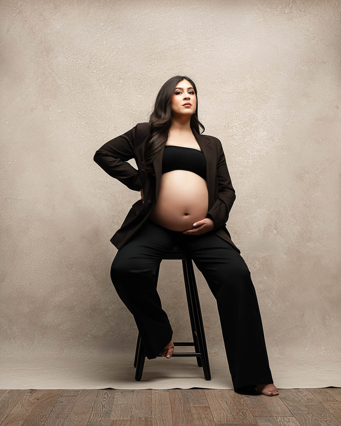 Editorial studio maternity photography in Dallas featuring an expecting mother seated in a black blazer and pants, showing her baby bump against a neutral textured backdrop