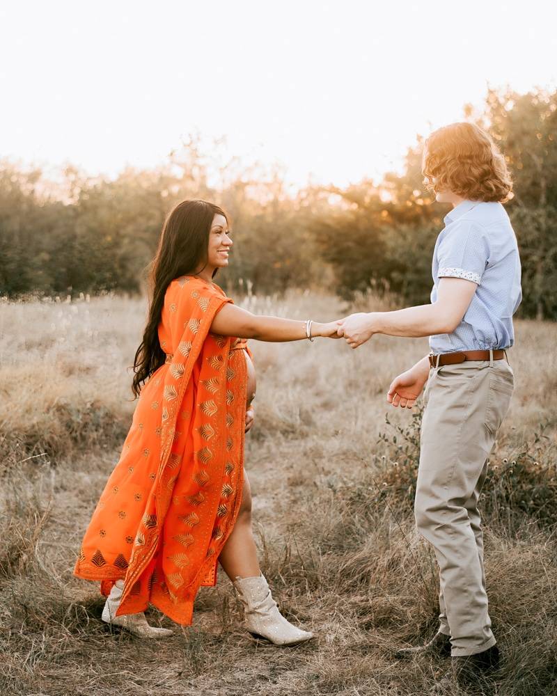 couple walks hand in hand through a grassy field smiling at camera for pregnancy photographer dallas