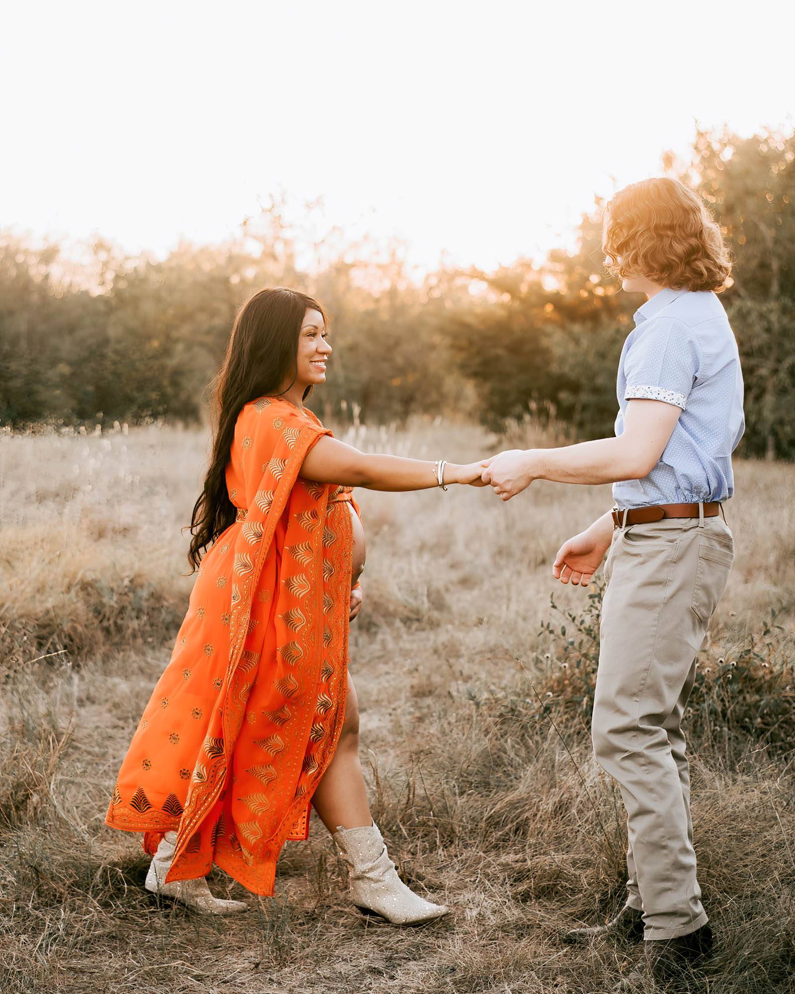 couple walks hand in hand through a grassy field smiling at camera for pregnancy photographer dallas
