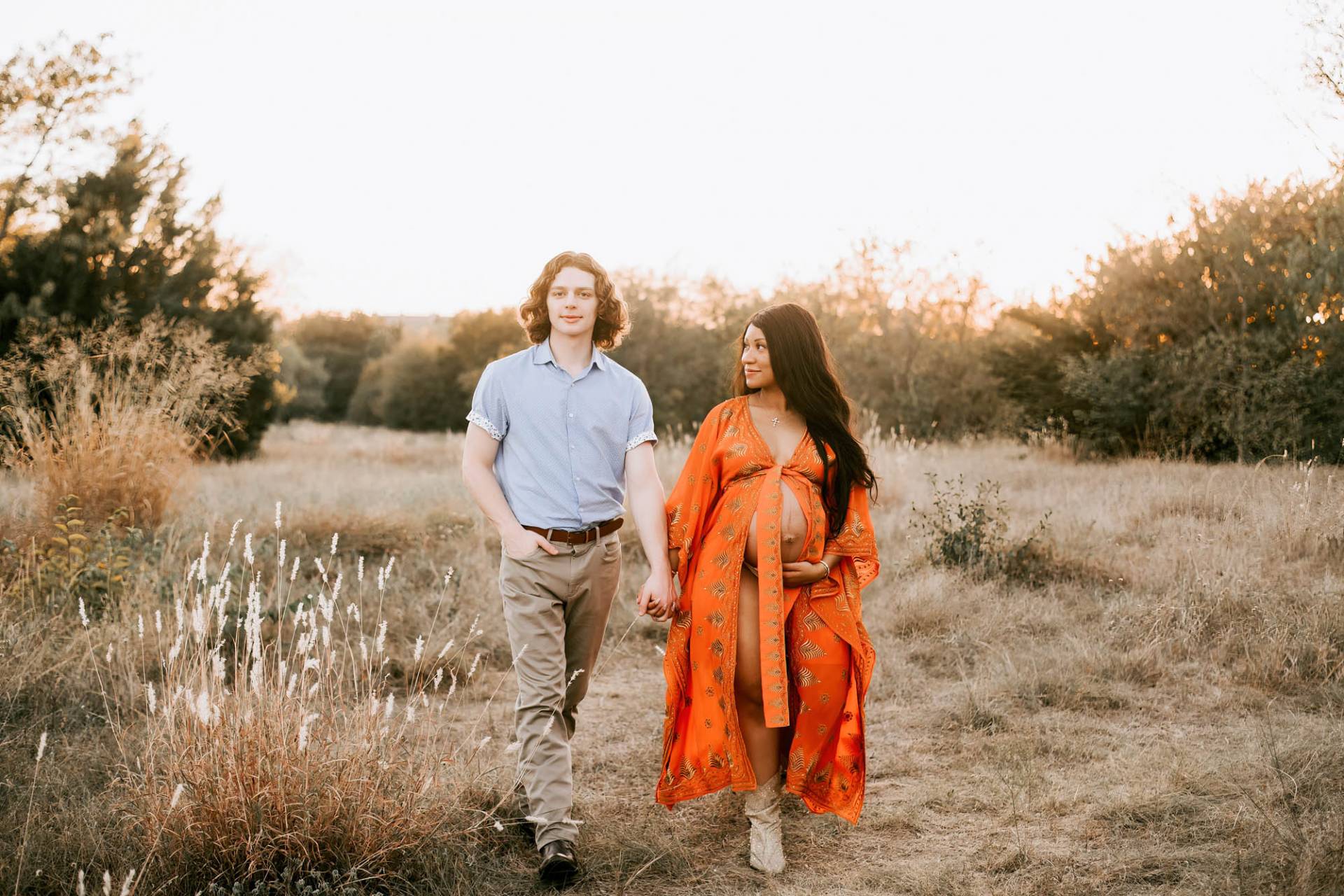 couple walks hand in hand through a grassy field smiling at camera for pregnancy photographer dallas